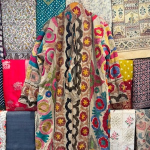 May include: A long-sleeved beige coat featuring vibrant, multi-colored floral and circular embroidery patterns. The garment includes a central vertical black wavy trim, a tie-front closure, and a structured collar, showcasing intricate needlework.