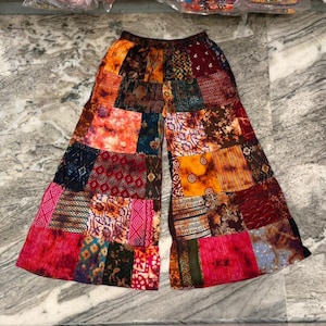 May include: Patchwork trousers in a wide-leg style. The fabric is composed of various patterned squares in red, orange, blue, and brown hues. The trousers are laid flat, displaying the full width and design of the fabric.