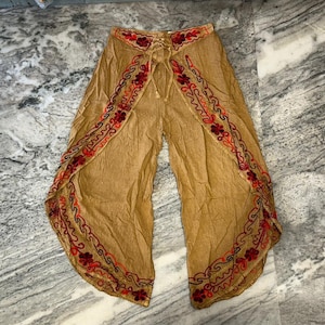 Puede incluir: Pantalones color arena con diseño envolvente. Bordados intrincados en rojo, naranja y dorado adornan los bordes y la cintura. La tela ligera sugiere un estilo bohemio o casual. Ideales para un look veraniego.