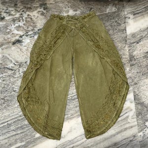 Könnte beinhalten: Olivgrüne Palazzo-Hose mit Spitzenbesatz an den Kanten. Die Hose hat einen Kordelzug in der Taille und ein weites Bein. Der Stoff scheint leicht und für warmes Wetter geeignet zu sein.