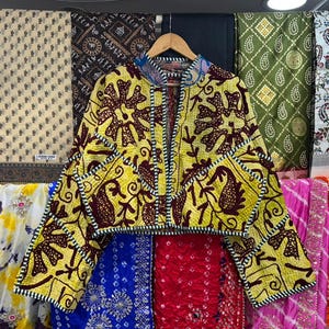 May include: A yellow jacket with a brown floral pattern and a blue collar. The jacket is hanging on a wooden hanger in front of a backdrop of patterned fabrics. The jacket has a cropped design and long sleeves.