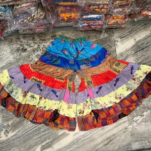 Może przedstawiać: Kolorowa spódnica patchwork z szerokim, rozkloszowanym krojem. Spódnica wykonana jest z różnych tkanin w odcieniach niebieskiego, pomarańczowego, czerwonego, fioletowego i żółtego. Niektóre panele zdobią motywy ptaków. Spódnica leży na marmurowej powierzchni.