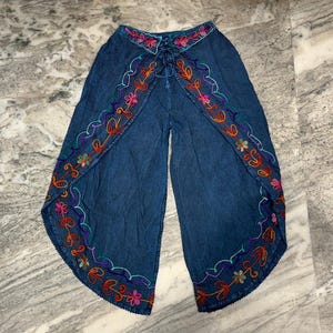May include: Blue palazzo pants with colorful embroidered floral and leaf designs along the edges. The pants have a wrap-style front with a drawstring closure at the waist. The embroidery features shades of orange, pink, green, and purple.