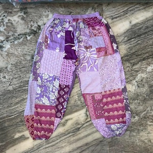 May include: Loose-fitting trousers featuring a patchwork design in various shades of purple and lavender. The garment displays a mix of floral, paisley, and geometric patterns across the fabric, with an elasticated waistband and gathered cuffs.