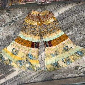 Op de afbeelding: Wijde broek met een gelaagd, uitlopend silhouet en een elastische tailleband. Het kledingstuk heeft een patchworkontwerp met diverse patronen in tinten geel, goud, bruin en lichtblauw, voorzien van paisley-motieven en horizontale banden.