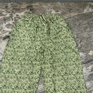 May include: Green patterned trousers with a drawstring waist. The trousers feature a repeating pattern of white and green floral and paisley designs. The fabric appears to be lightweight and suitable for warm weather.