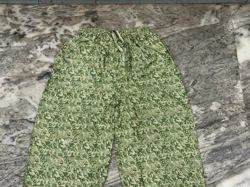 May include: Green patterned trousers with a drawstring waist. The trousers feature a repeating pattern of white and green floral and paisley designs. The fabric appears to be lightweight and suitable for warm weather.