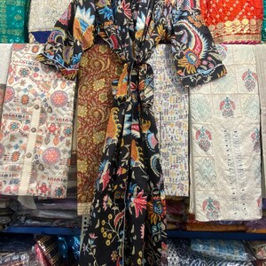May include: A black kimono robe with a colourful floral and paisley design in red, blue, yellow, and green. The robe is displayed on a wooden hanger, with a matching tie belt. The background shows various textiles.