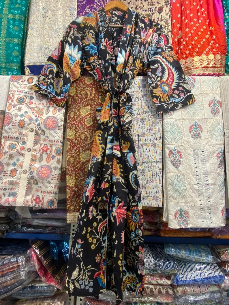 May include: A black kimono robe with a colourful floral and paisley design in red, blue, yellow, and green. The robe is displayed on a wooden hanger, with a matching tie belt. The background shows various textiles.