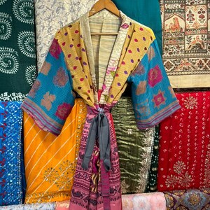 May include: A colorful kimono-style robe with a tie belt, displayed on a wooden hanger. The robe features a patchwork design with blue, yellow, and pink floral patterns. Various patterned fabrics in shades of blue, green, yellow, and red are visible in the background.