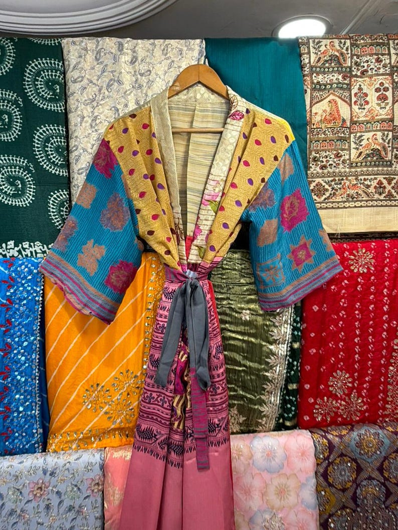 May include: A colorful kimono-style robe with a tie belt, displayed on a wooden hanger. The robe features a patchwork design with blue, yellow, and pink floral patterns. Various patterned fabrics in shades of blue, green, yellow, and red are visible in the background.