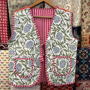 May include: A reversible quilted vest with a white background and a floral pattern of blue fruit and green leaves. The vest has a pink striped lining and trim, with orange accents on the shoulders and pockets.