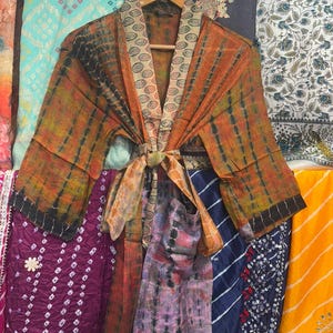May include: A tie-dye kimono in shades of orange and black, with a patterned collar and tie. The robe is displayed on a wooden hanger, with colourful fabrics behind it. The robe features long sleeves and a tie closure.