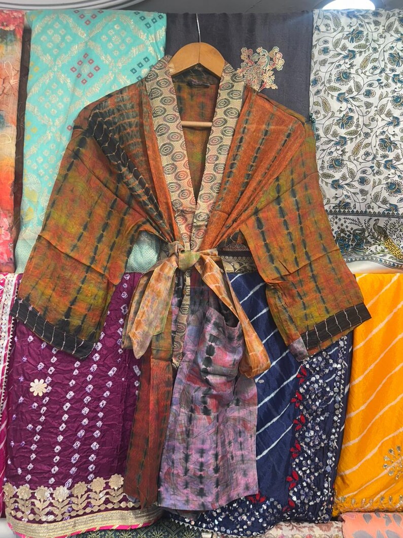 May include: A tie-dye kimono in shades of orange and black, with a patterned collar and tie. The robe is displayed on a wooden hanger, with colourful fabrics behind it. The robe features long sleeves and a tie closure.