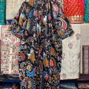 May include: A black floral print robe with white edging, hanging on a wooden hanger. The robe displays a colourful pattern of flowers and birds in red, blue, yellow, and pink. The robe is shown against a backdrop of various patterned fabrics.