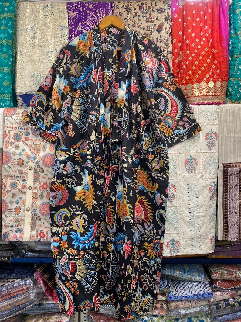May include: A black floral print robe with white edging, hanging on a wooden hanger. The robe displays a colourful pattern of flowers and birds in red, blue, yellow, and pink. The robe is shown against a backdrop of various patterned fabrics.