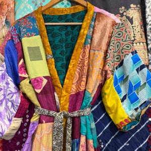 May include: A patchwork silk kimono robe with a vibrant mix of colors and patterns, including floral, paisley, and geometric designs. The robe features a mustard yellow trim and a matching belt. The robe is hanging on a wooden hanger.