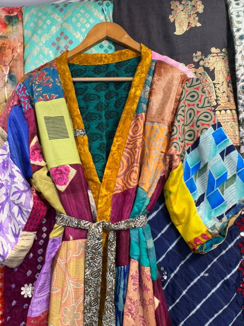 May include: A patchwork silk kimono robe with a vibrant mix of colors and patterns, including floral, paisley, and geometric designs. The robe features a mustard yellow trim and a matching belt. The robe is hanging on a wooden hanger.