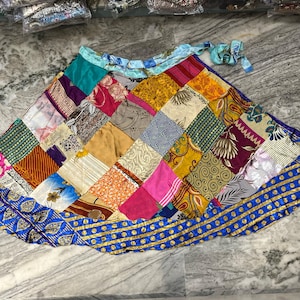 Puede incluir: Una falda envolvente de patchwork colorida hecha de saris reciclados. La falda es azul, rosa, verde, amarilla y marrón. Tiene un cierre de nudo y un ribete dorado.