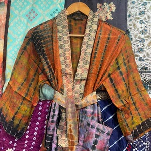 May include: A vibrant, tie-dye patterned kimono robe in shades of orange, black, and yellow, with a matching belt. The robe is displayed on a wooden hanger, highlighting its flowing design and bright colours. The background features various patterned fabrics.