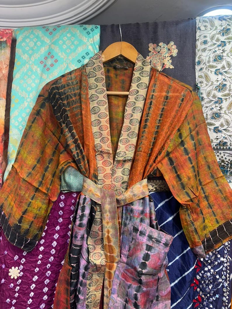 May include: A vibrant, tie-dye patterned kimono robe in shades of orange, black, and yellow, with a matching belt. The robe is displayed on a wooden hanger, highlighting its flowing design and bright colours. The background features various patterned fabrics.