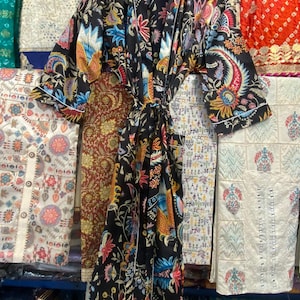 May include: A black floral print dressing gown with a tie belt and white edging. The gown has a colourful pattern of flowers, birds, and paisley designs. The garment is hanging on a wooden hanger, with other patterned fabrics in the background.