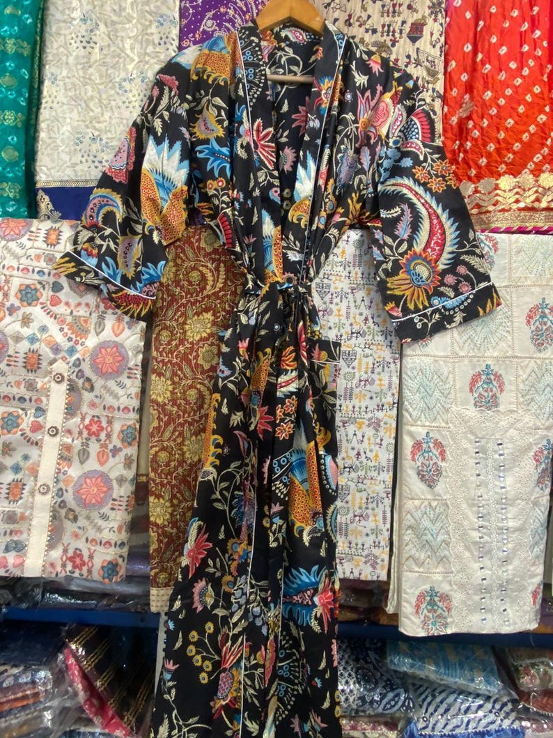 May include: A black floral print dressing gown with a tie belt and white edging. The gown has a colourful pattern of flowers, birds, and paisley designs. The garment is hanging on a wooden hanger, with other patterned fabrics in the background.