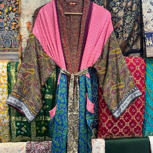 Peut inclure: Une robe kimono en soie colorée avec un motif cachemire. La robe présente un corsage rose, des manches cachemire et une section inférieure à motifs bleus et verts. Elle possède une ceinture et des poches assorties. La robe est suspendue à un cintre en bois.