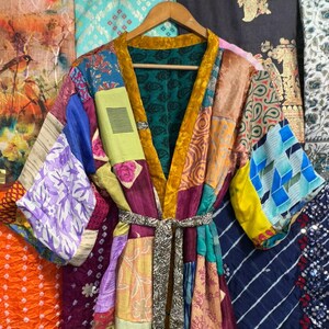 May include: A patchwork kimono robe with a variety of colorful fabric patterns. The robe has a gold velvet trim and a matching belt. The sleeves and body are made of different fabrics, including floral and geometric designs. The robe is hanging on a wooden hanger.