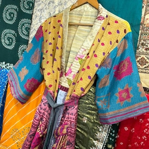 May include: A colorful, patchwork kimono robe with a gray tie belt, hanging on a wooden hanger. The robe features a mix of patterns and colors, including yellow with purple dots, blue floral sleeves, and pink and red patterned panels.