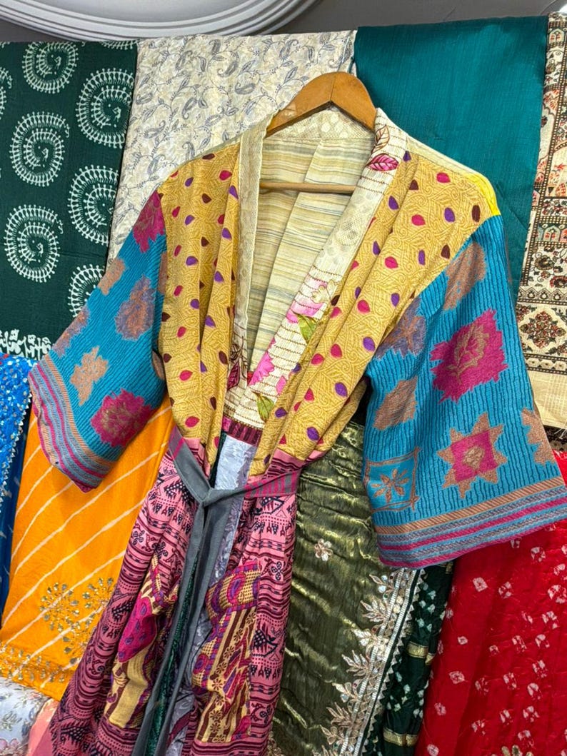 May include: A colorful, patchwork kimono robe with a gray tie belt, hanging on a wooden hanger. The robe features a mix of patterns and colors, including yellow with purple dots, blue floral sleeves, and pink and red patterned panels.