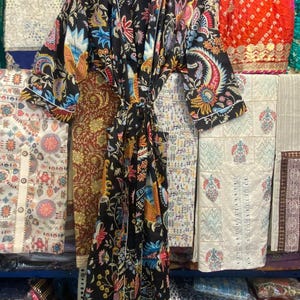 May include: A black patterned robe with a vibrant floral design in shades of blue, orange, and pink. The robe has white edging and a tie. It is displayed on a wooden hanger, highlighting the garment's intricate pattern and style.