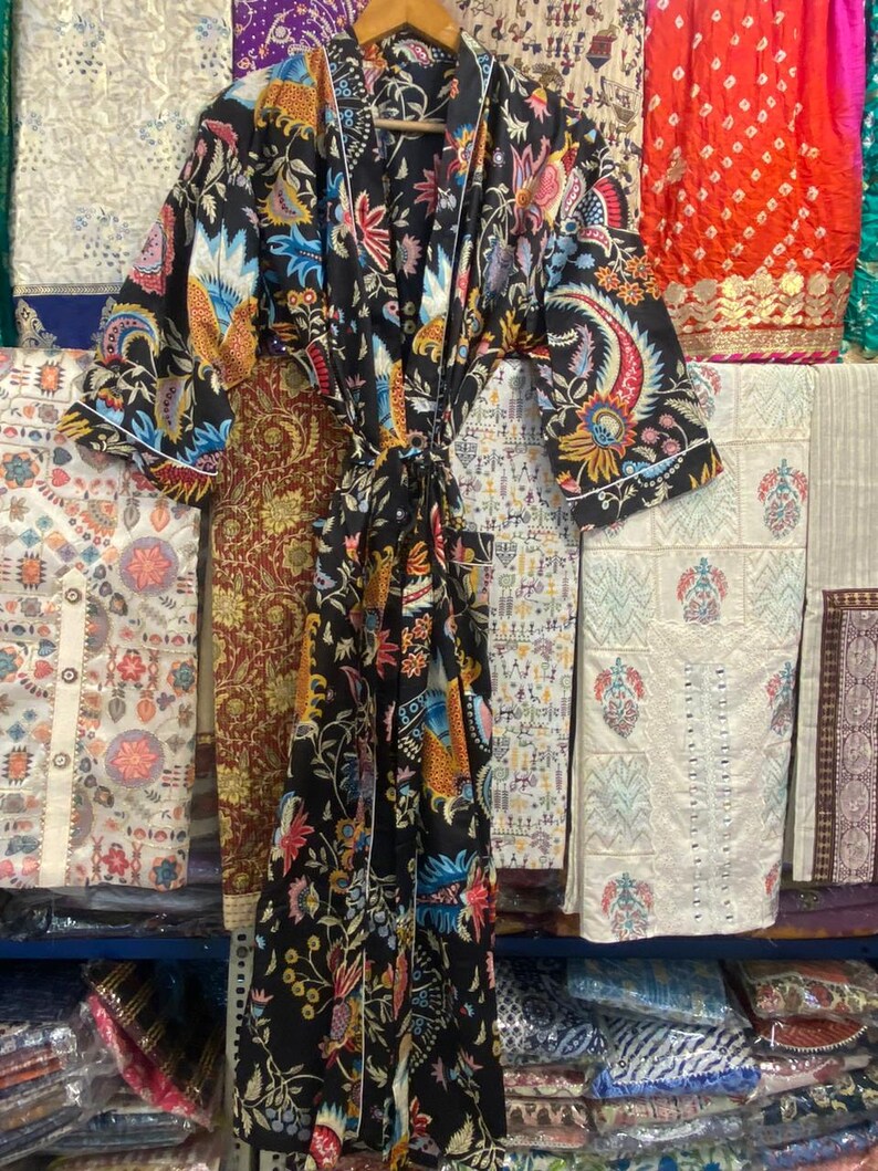 May include: A black patterned robe with a vibrant floral design in shades of blue, orange, and pink. The robe has white edging and a tie. It is displayed on a wooden hanger, highlighting the garment's intricate pattern and style.