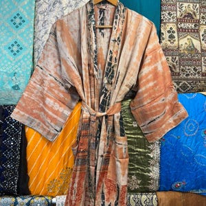 May include: A long-sleeved robe featuring a tie-waist belt and two front pockets. The garment displays a tie-dye pattern in shades of peach, orange, and dark charcoal gray, with a contrasting vertical panel design along the front opening.