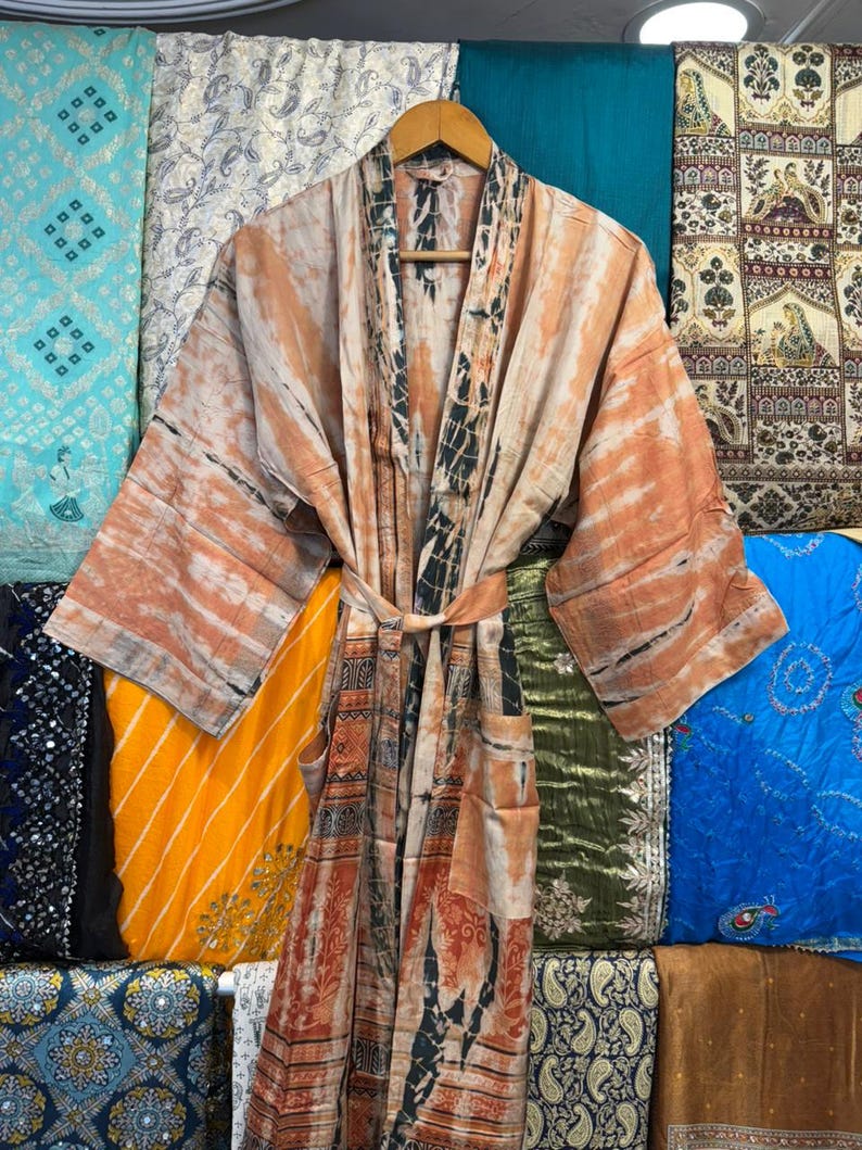 May include: A long-sleeved robe featuring a tie-waist belt and two front pockets. The garment displays a tie-dye pattern in shades of peach, orange, and dark charcoal gray, with a contrasting vertical panel design along the front opening.