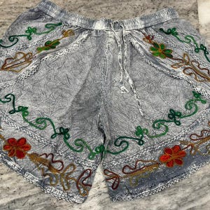May include: Gray shorts with an elastic waistband and drawstring. The shorts are decorated with embroidered floral patterns in red, green, and brown. The shorts have a textured appearance.