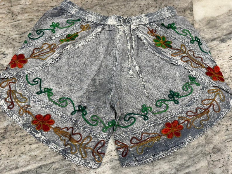May include: Gray shorts with an elastic waistband and drawstring. The shorts are decorated with embroidered floral patterns in red, green, and brown. The shorts have a textured appearance.