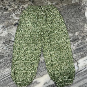 May include: Green patterned harem trousers with an elasticated waistband and drawstring. The trousers feature a repeating floral design in shades of green and white. The loose-fitting trousers have a gathered ankle.