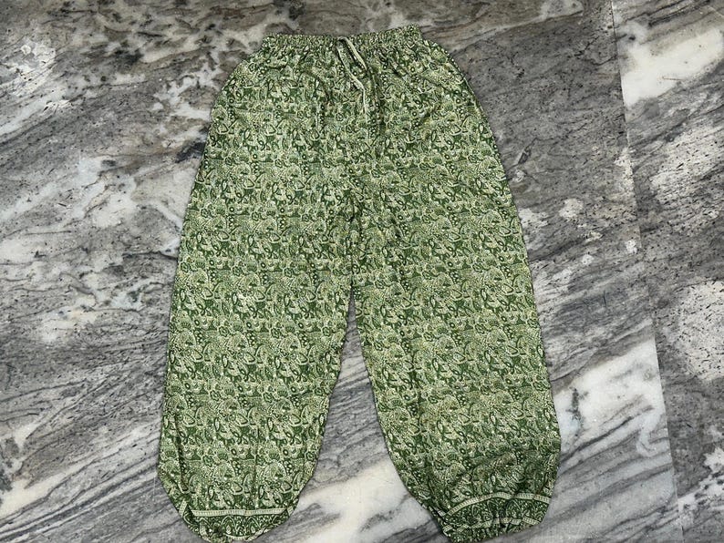 May include: Green patterned harem trousers with an elasticated waistband and drawstring. The trousers feature a repeating floral design in shades of green and white. The loose-fitting trousers have a gathered ankle.