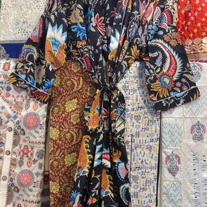 May include: A black kimono-style robe with a vibrant floral pattern in shades of red, blue, yellow, and orange. The robe is tied at the waist with a matching fabric belt. The sleeves are wide and the robe is hanging on a wooden hanger.