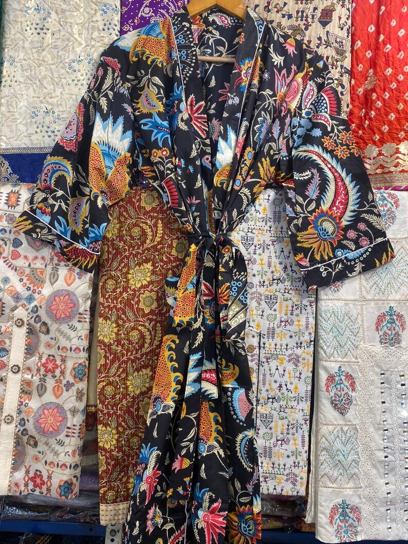 May include: A black kimono-style robe with a vibrant floral pattern in shades of red, blue, yellow, and orange. The robe is tied at the waist with a matching fabric belt. The sleeves are wide and the robe is hanging on a wooden hanger.
