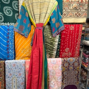 May include: A colorful, patterned robe with a matching belt hangs on a wooden hanger. The robe features blue, yellow, and red panels, with patterned sleeves. The background displays various fabrics in different colors and patterns.