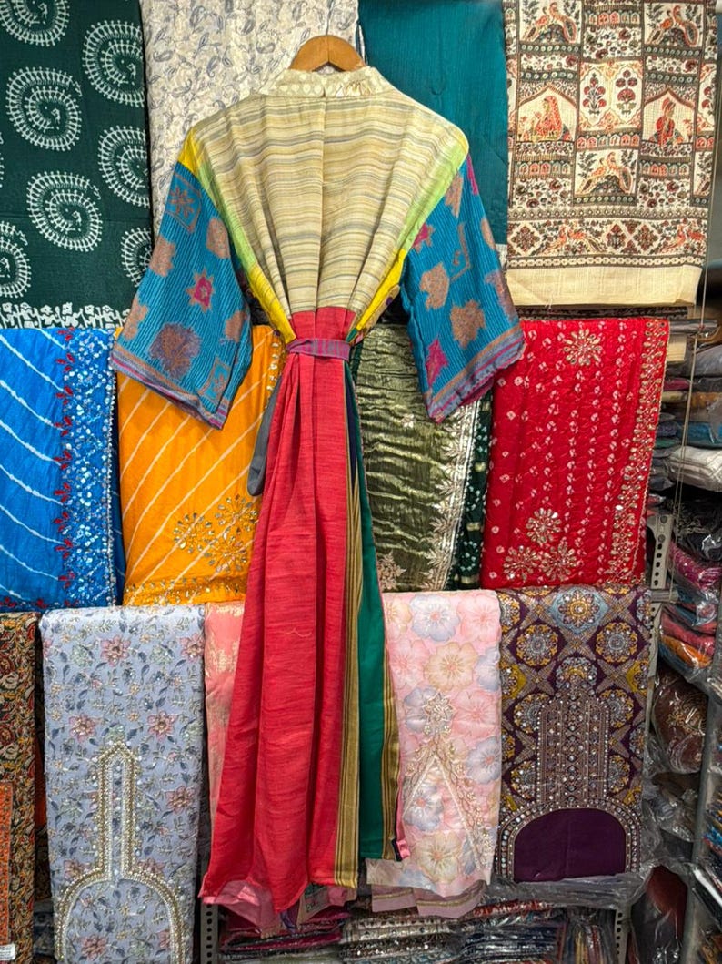 May include: A colorful, patterned robe with a matching belt hangs on a wooden hanger. The robe features blue, yellow, and red panels, with patterned sleeves. The background displays various fabrics in different colors and patterns.