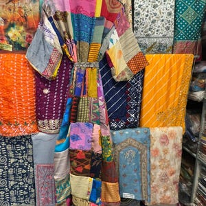 May include: A patchwork kimono robe with a variety of colorful fabric patterns, hanging on a wooden hanger. The robe is made of different fabrics and colors, including pink, blue, yellow, and purple. The background features various textiles.