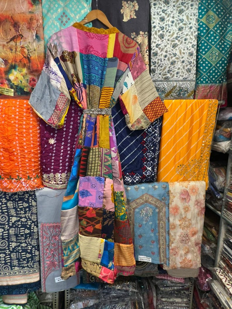 May include: A patchwork kimono robe with a variety of colorful fabric patterns, hanging on a wooden hanger. The robe is made of different fabrics and colors, including pink, blue, yellow, and purple. The background features various textiles.
