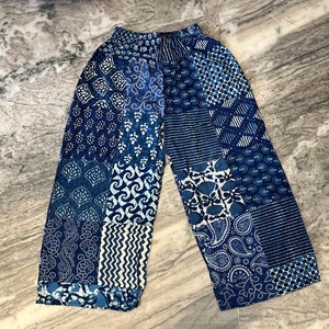Op de afbeelding: Blauw-witte patchworkbroek met diverse geometrische en bloemmotieven. De broek heeft een elastische tailleband en een wijde pijp. De stof lijkt lichtgewicht, geschikt voor warm weer.