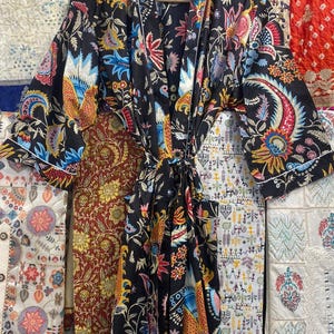 May include: A black patterned robe with a vibrant floral design in shades of blue, red, and yellow. The robe features long sleeves and a white trim. It is displayed on a wooden hanger.