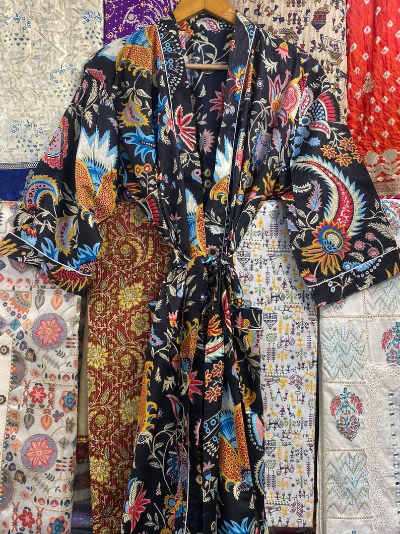 May include: A black patterned robe with a vibrant floral design in shades of blue, red, and yellow. The robe features long sleeves and a white trim. It is displayed on a wooden hanger.