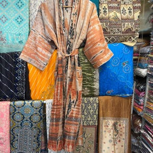 May include: A long-sleeved robe featuring a tie-waist belt and a tie-dye pattern in shades of orange, brown, and cream. The garment has a wide collar and two front pockets, displayed on a wooden hanger against a backdrop of various folded fabrics.