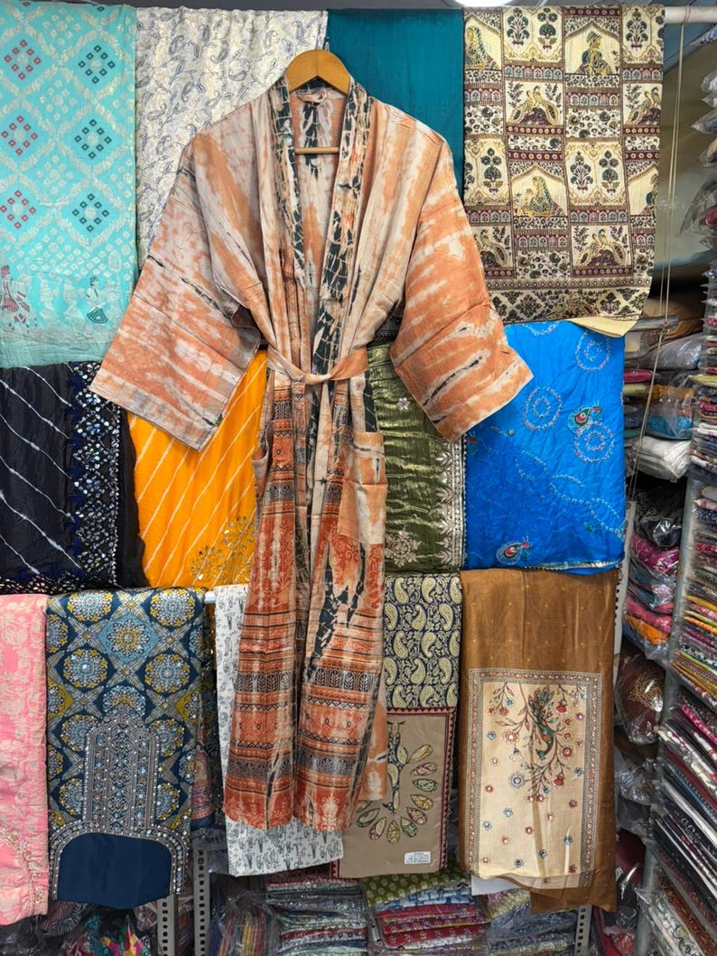 May include: A long-sleeved robe featuring a tie-waist belt and a tie-dye pattern in shades of orange, brown, and cream. The garment has a wide collar and two front pockets, displayed on a wooden hanger against a backdrop of various folded fabrics.
